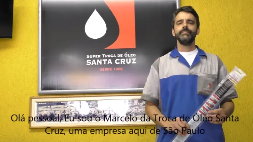 Um homem de uniforme azul e cinza segura um refil de palheta, diante do letreiro da Super Troca de Óleo Santa Cruz.